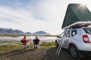 Young couple sitting in folding chairs next to offroad car with tent on the roof.